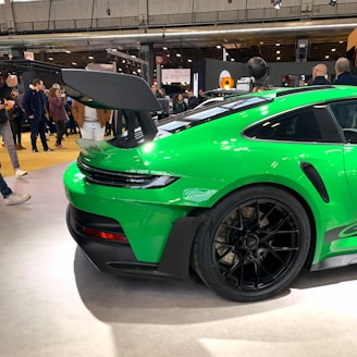 A vibrant green sports car is prominently displayed indoors, showcasing its sleek design and large rear spoiler. The car is surrounded by people, likely at a car exhibition or showroom, emphasizing its high-performance features and appealing aesthetics.