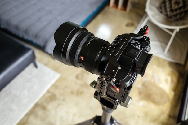 A professional camera with an attached lens mounted on a tripod is positioned indoors. The setting includes a modern interior with a bed, chair, and decorative items visible in the blurred background.