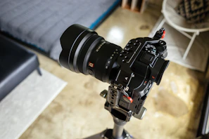 A professional camera with an attached lens mounted on a tripod is positioned indoors. The setting includes a modern interior with a bed, chair, and decorative items visible in the blurred background.