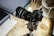 A professional camera with an attached lens mounted on a tripod is positioned indoors. The setting includes a modern interior with a bed, chair, and decorative items visible in the blurred background.