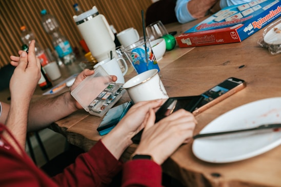 A wooden table with people gathered around, engaging in an activity involving a board game and various objects. The table holds mugs, a bottle, a plate, a smartphone, and a game box labeled 'Rummikub'. One person is holding a case with what appears to be game pieces or cards, while another is interacting with a smartphone.