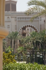 A scenic view of a Middle Eastern architectural building with ornate details, surrounded by palm trees and greenery. Traditional lanterns line the foreground, contributing to the elegance and cultural atmosphere of the setting.