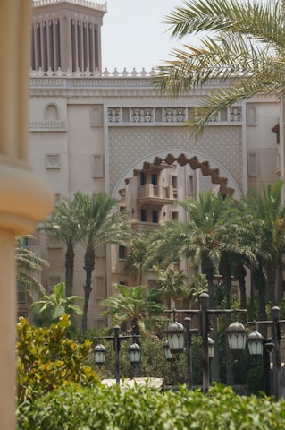 A scenic view of a Middle Eastern architectural building with ornate details, surrounded by palm trees and greenery. Traditional lanterns line the foreground, contributing to the elegance and cultural atmosphere of the setting.