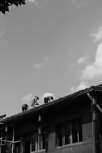 A black and white photograph showing three people working on the roof of a building. The structure appears to be made of wood with tiled roofing. The workers are positioned along the sloped roof, bent over, seemingly focused on their tasks. The sky above is partly cloudy, and some leafy branches are visible from the top left corner.