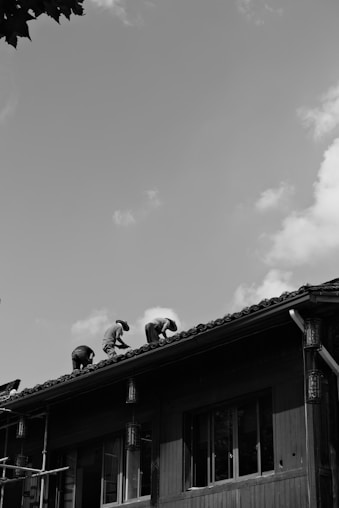 A black and white photograph showing three people working on the roof of a building. The structure appears to be made of wood with tiled roofing. The workers are positioned along the sloped roof, bent over, seemingly focused on their tasks. The sky above is partly cloudy, and some leafy branches are visible from the top left corner.