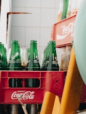 A collection of green Sprite bottles is arranged in a red Coca-Cola crate. The crate is positioned against a tiled wall, partially obscured by other objects. The bottles are empty, and the setup suggests a storage or recycling area.
