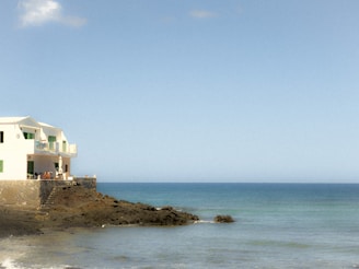 A white building with green shutters is situated on a rocky shoreline, overlooking a calm sea under a clear blue sky. A couple of people are visible on the terrace, enjoying the seaside view.