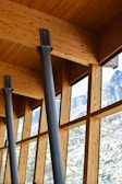 Large wooden beams and metal columns form part of an architectural structure with a series of tall windows overlooking a mountainous landscape. The interior features light wood grain, while the windows offer a view of snow-capped peaks and rocky terrain.