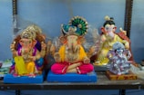 Four colorful statues of the Hindu deity Ganesha are displayed on a shelf. The statues vary in design, with intricate decorations and vibrant colors. Each figure is depicted with traditional elements such as jewels, clothing, and paint details, exhibiting craftsmanship in their structure.