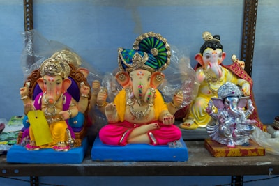 Four colorful statues of the Hindu deity Ganesha are displayed on a shelf. The statues vary in design, with intricate decorations and vibrant colors. Each figure is depicted with traditional elements such as jewels, clothing, and paint details, exhibiting craftsmanship in their structure.