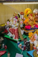 A collection of intricately designed and colorful figurines depicting Hindu deities, prominently featuring Ganesha. The statues are adorned with ornate headpieces, vibrant clothing, and jewelry. At the front, art supplies like paint bottles, brushes, and fabric pieces are visible, suggesting an ongoing creative process.
