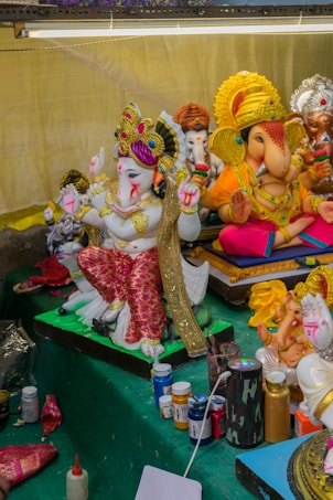 A collection of intricately designed and colorful figurines depicting Hindu deities, prominently featuring Ganesha. The statues are adorned with ornate headpieces, vibrant clothing, and jewelry. At the front, art supplies like paint bottles, brushes, and fabric pieces are visible, suggesting an ongoing creative process.