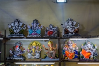 The image features multiple statues of the Hindu deity Ganesha placed on metal shelves. The statues are made of different materials and are decorated with various colors like gold, blue, orange, yellow, and red. Each Ganesha statue is distinctly crafted, some holding objects while others are in different poses. The background is plain, and the area is illuminated with bright lighting.