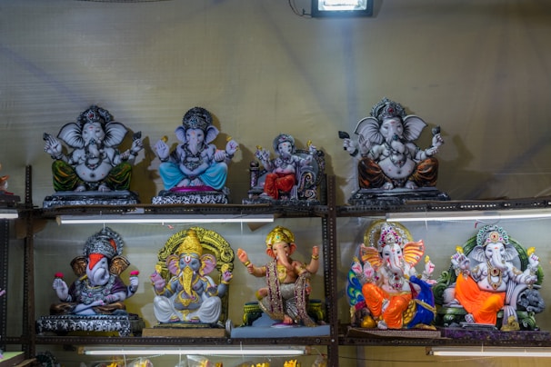 The image features multiple statues of the Hindu deity Ganesha placed on metal shelves. The statues are made of different materials and are decorated with various colors like gold, blue, orange, yellow, and red. Each Ganesha statue is distinctly crafted, some holding objects while others are in different poses. The background is plain, and the area is illuminated with bright lighting.