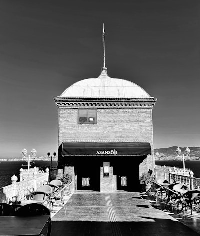 A monochrome image depicting a building with a dome and a spire, labeled 'Asansör'. The structure is situated near a large body of water with a view of distant cityscape. Wrought-iron railings and street lamps are visible, along with several empty patio chairs and tables.