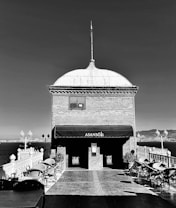 A monochrome image depicting a building with a dome and a spire, labeled 'Asans&ouml;r'. The structure is situated near a large body of water with a view of distant cityscape. Wrought-iron railings and street lamps are visible, along with several empty patio chairs and tables.