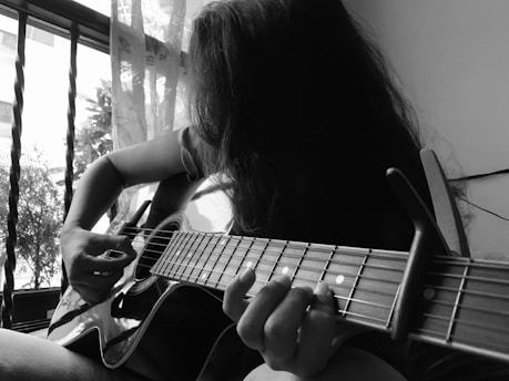 A person plays an acoustic guitar near a window with bars and partially drawn curtains. The focus is on their hand strumming the strings and the guitar body. The scene is in black and white, adding a serene and introspective feel.