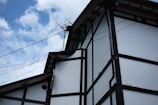 A traditional-style building with white walls and dark wooden beams is depicted against a partly cloudy sky. The roof features multiple angled sections, and there are antennas mounted on top. Electrical wires are visible, running across the building.