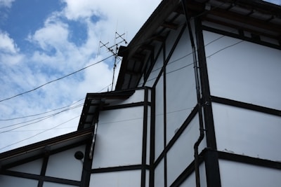 A traditional-style building with white walls and dark wooden beams is depicted against a partly cloudy sky. The roof features multiple angled sections, and there are antennas mounted on top. Electrical wires are visible, running across the building.