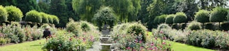 A beautifully manicured garden with neatly trimmed hedges and vibrant flower beds. A serene water feature runs through the center, and a person wearing a hat sits among the blooms, enjoying the peaceful environment. Tall trees surround the garden, providing shade and a sense of seclusion.