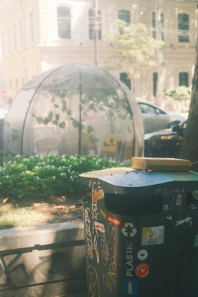 An outdoor scene showing a trash can adorned with various stickers in the foreground. Behind the trash can, there is a dome-shaped greenhouse or plant enclosure with visible green plants inside. There are buildings and parked cars in the background, and flowers and shrubs in the middle ground.