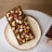 A rectangular chocolate bar is covered with various toppings, including cashews, pistachios, dried fruit pieces, golden edible flakes, and other nuts. The bar is placed on a round ceramic plate, which is set on a white surface. There are also parts of a magazine or book visible in the bottom right corner.