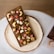 A rectangular chocolate bar is covered with various toppings, including cashews, pistachios, dried fruit pieces, golden edible flakes, and other nuts. The bar is placed on a round ceramic plate, which is set on a white surface. There are also parts of a magazine or book visible in the bottom right corner.