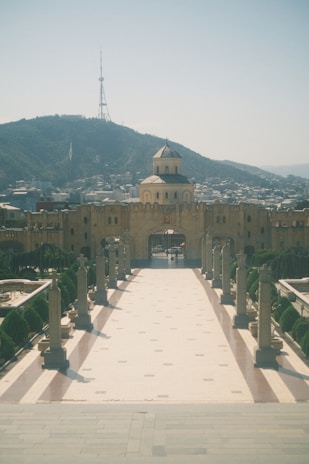 A broad pathway lined with columns leads to an ornate gate, behind which stands a domed building. The scene is framed by a distant mountainous backdrop that supports a tall communications tower.
