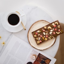 A cup of black coffee sits on a white saucer with a gold spoon, placed on a marble surface. Next to it is a magazine with text and a photograph of a hat. A plate holds a chocolate bar topped with various dried fruits and nuts, including cashews and pistachios.