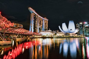 A stunning night view of a modern cityscape featuring an iconic hotel with three towers and a unique boat-shaped structure on top, brightly lit with colorful lights. A nearby white, futuristic building with a lotus-like design adds distinct architectural flair. The red-lit bridge is reflected in the calm waters, enhancing the vibrant scene.