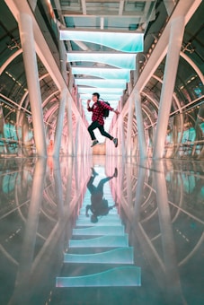A person is jumping energetically in a futuristic hallway, which features an array of illuminated, turquoise-colored panels on the ceiling and a shiny, reflective floor. The structure of the hall is arch-shaped with large metallic supports.