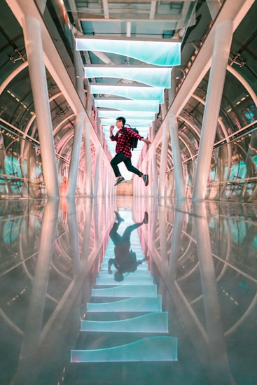 A person is jumping energetically in a futuristic hallway, which features an array of illuminated, turquoise-colored panels on the ceiling and a shiny, reflective floor. The structure of the hall is arch-shaped with large metallic supports.
