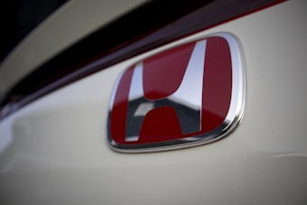 A close-up of the Honda logo on a vehicle. The iconic emblem features a white letter 'H' against a red background, surrounded by a shiny silver or chrome frame. The surrounding area is a smooth, creamy white surface, possibly the body of a car.