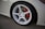 A close-up view of a car wheel featuring a white multi-spoke alloy rim. The tire is mounted on a car with a light-colored body, and a red brake caliper is visible through the spokes.