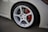 A close-up view of a car wheel featuring a white multi-spoke alloy rim. The tire is mounted on a car with a light-colored body, and a red brake caliper is visible through the spokes.