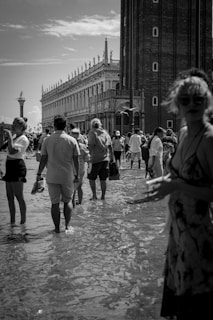 A black and white photograph of people walking through shallow water in a city square, with historical architecture and a bell tower in the background. The mood is bustling with several individuals capturing photos on their phones. A seagull is flying near the ornate building with an overcast sky above.