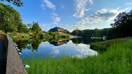 A serene lake with crystal-clear reflections of lush green trees and a building in the distance, under a sky dotted with fluffy clouds. A wooden walkway borders the left side of the image, and vibrant greenery frames the foreground.