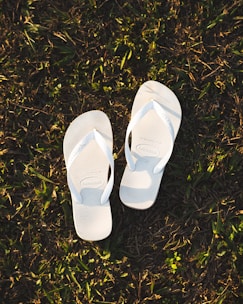 A pair of white flip-flops is placed on grass, casting soft shadows due to the warm sunlight.
