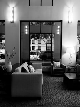 A modern living room featuring a large sofa with patterned cushions, an armchair, a coffee table, and a decorative vase on a side table. Tall windows reveal a street view with a parked car and a bicycle. Interior lights add a warm ambiance.