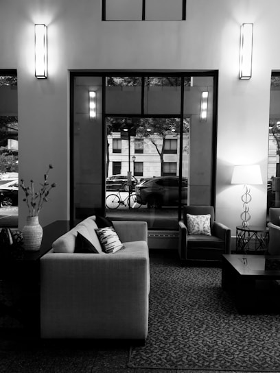 A modern living room featuring a large sofa with patterned cushions, an armchair, a coffee table, and a decorative vase on a side table. Tall windows reveal a street view with a parked car and a bicycle. Interior lights add a warm ambiance.