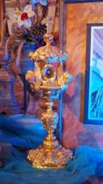 A gold ornate chalice with intricate details and a cross on top is centrally placed. Surrounding it are decorative elements including flowers and blue fabric, creating a rich, ceremonial atmosphere. An ornate frame with artwork is partially visible in the background.