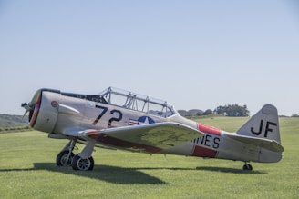 A vintage military aircraft with a metal fuselage parked on a grassy airfield. The aircraft is painted in a light gray color with the number 72 and military insignia on its side. The background features a clear blue sky and some distant trees.