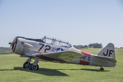 A vintage military aircraft with a metal fuselage parked on a grassy airfield. The aircraft is painted in a light gray color with the number 72 and military insignia on its side. The background features a clear blue sky and some distant trees.