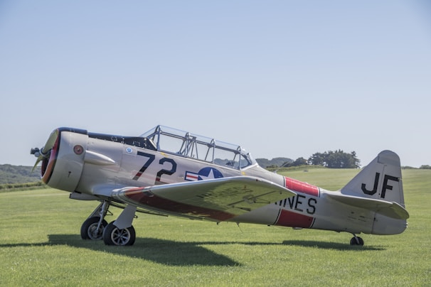 A vintage military aircraft with a metal fuselage parked on a grassy airfield. The aircraft is painted in a light gray color with the number 72 and military insignia on its side. The background features a clear blue sky and some distant trees.