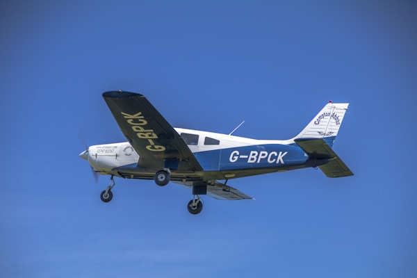 A small aircraft with the registration G-BPCK is flying against a clear blue sky. The airplane is primarily white with blue accents and visible branding on the side and tail. The landing gear is extended.