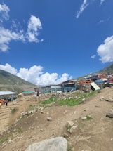 A rural landscape featuring a dirt path leading through a small settlement of makeshift structures surrounded by rocky terrain. A few people are visible walking along the path, and the sky above is bright blue with scattered white clouds. Green hills flank the scene, providing a backdrop to the modest buildings constructed with corrugated metal and wood.