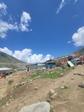 A rural landscape featuring a dirt path leading through a small settlement of makeshift structures surrounded by rocky terrain. A few people are visible walking along the path, and the sky above is bright blue with scattered white clouds. Green hills flank the scene, providing a backdrop to the modest buildings constructed with corrugated metal and wood.