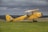 A vintage yellow biplane with British roundels is parked on a grassy field, set against a backdrop of cloudy skies and distant rolling hills. The plane's design suggests a historical model. A person wearing a helmet is seated in the cockpit.