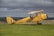 A vintage yellow biplane with British roundels is parked on a grassy field, set against a backdrop of cloudy skies and distant rolling hills. The plane's design suggests a historical model. A person wearing a helmet is seated in the cockpit.