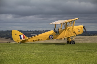 A vintage yellow biplane with British roundels is parked on a grassy field, set against a backdrop of cloudy skies and distant rolling hills. The plane's design suggests a historical model. A person wearing a helmet is seated in the cockpit.